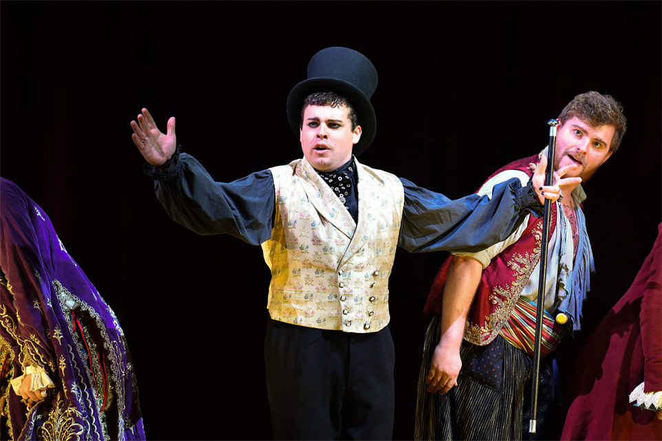 A male student, wearing a yellow waistcoat, top hat and holding a cane, singing to the audience, with the chorus singing beside him.